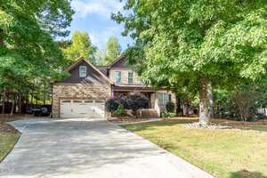 A two-story house with stone and brown siding, a two-car garage, front porch, and an American flag. The house is surrounded by trees and has a concrete driveway leading to the street.
