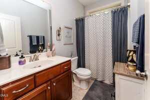 Bathroom with a wooden vanity, white sink, and marble countertop. Toilet next to a small cabinet with decor. Gray walls, a shower with a white and gray curtain, towel racks, and a wall sign complete the room.