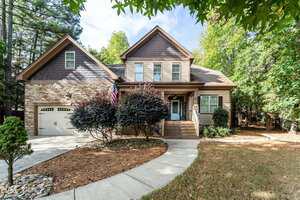 Two-story suburban house with stone and wood exterior, a covered front porch, and attached two-car garage. Concrete driveway curves toward the entrance, with landscaped shrubs and trees surrounding the lawn.