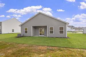 A single-story gray house with a sloped roof, three windows, and double glass doors opening to a grassy backyard. An air conditioning unit is installed on the left side. Other houses are visible in the background.