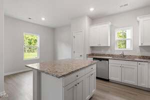 A modern kitchen with white cabinets, granite countertops, a central island, stainless steel dishwasher, and light wood flooring. Two windows provide natural light and a door is visible in the background.