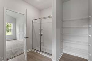 A bathroom with a glass-enclosed shower, white walls, wood-look flooring, and white wire shelving. An open door leads to a carpeted room with a window.