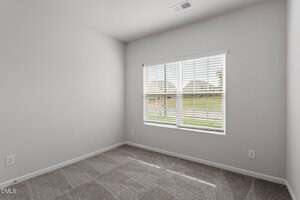 Empty room with light gray carpet, white walls, two electrical outlets, and a large window with white blinds letting in natural light. The window shows a grassy outdoor area and some fencing.