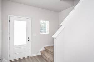 A bright entryway with a white front door, a small window with blinds, carpeted stairs, and light-colored walls and flooring.