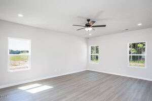 A spacious, empty room with light gray wood flooring, white walls, three windows with outdoor views, and a black ceiling fan with a light fixture in the center of the ceiling.