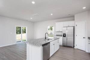 Modern kitchen with white cabinets, stainless steel appliances, granite countertops, and an island with a sink. There is a dishwasher and a window above the counter. Sliding glass doors lead to a backyard. Light wood flooring throughout.
