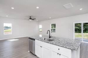 A modern open-concept kitchen and living area with light gray wood flooring, white walls, a granite countertop island with sink and dishwasher, and several windows letting in natural light.