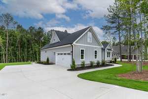 A light gray single-story house with white trim and a steep roof sits beside a large, curved concrete driveway. The yard is landscaped with green grass, small shrubs, and surrounded by tall trees.