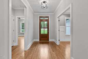 A hallway with light gray walls, wooden floors, and a wooden front door with glass panels; rooms with windows and doors are visible on both sides, and a ceiling light fixture is above.