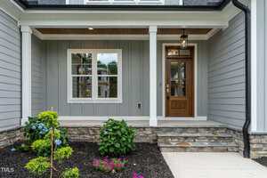 A modern house entryway with gray siding, a wooden front door with glass panels, white trim, a stone porch, two columns, and a small landscaped garden with shrubs and flowers.