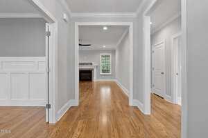 A hallway with polished wood floors, light gray walls, white trim, and several doorways leading to different rooms. A fireplace and large window are visible in the background living area. Ceiling lights are turned on.