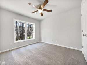 A vacant room with beige carpet, light gray walls, a ceiling fan with light, and a window with black trim providing a view of trees outside. Two closed white doors are visible on the right.