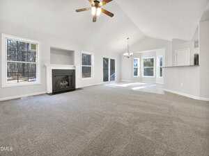 A spacious, empty living room with light gray carpet, white walls, a ceiling fan with lights, a fireplace, large windows, and an adjacent dining area with a bay window and overhead chandelier.