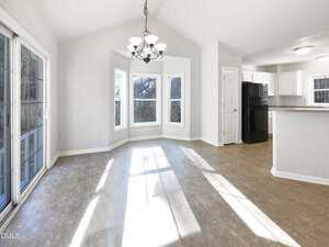 A bright, empty room with large windows, a hanging light fixture, tiled flooring, and an open view into a kitchen with white cabinets and black appliances. Sunlight streams across the floor.