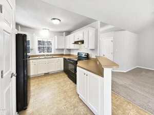 A kitchen with white cabinets, beige countertops, a stainless steel stove, black refrigerator, and tiled flooring. The space is open to a carpeted living area and has neutral-colored walls.