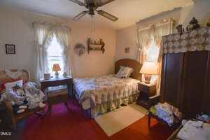 A cozy bedroom with a single bed, patchwork quilt, side tables with lamps, wicker chair with stuffed animals, red carpet, curtains on two windows, and various knick-knacks on shelves and furniture. Warm, inviting atmosphere.