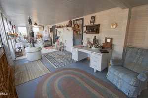 A cozy enclosed porch with wooden floors, area rugs, a white desk, blue armchair, various decorative items, and vintage furniture. The space is bright with natural light from windows along one wall.