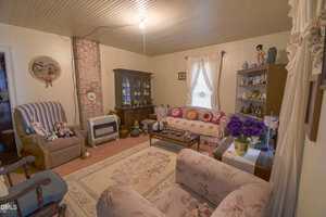 A cozy vintage living room with floral upholstered furniture, a brick-accented wall, a china cabinet, shelves with figurines, and a window with lacy curtains. Dolls and decorative items add a nostalgic touch.