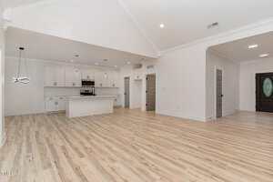 Spacious open-concept kitchen and living area with light wood floors, white walls, recessed lighting, and a central island. Modern cabinets and stainless appliances are visible; multiple doors lead to other rooms.