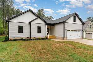 Single-story house with white vertical siding, black trim, and a two-car garage. The home has a small covered front entry, steps leading to the door, and a driveway, with grass and shrubs in the front yard.