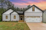 Single-story modern house at 1414 Sagamore Drive, Louisburg, with white siding, black trim, a double garage, and small front porch. The yard features a concrete driveway, grass, trees, and a colorful sky in the background.