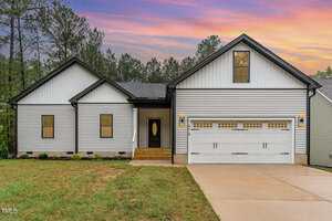 Single-story modern house at 1414 Sagamore Drive, Louisburg, with white siding, black trim, a double garage, and small front porch. The yard features a concrete driveway, grass, trees, and a colorful sky in the background.