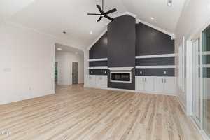 Spacious modern living room with light wood floors, a vaulted ceiling, a black ceiling fan, and a dark accent wall featuring a built-in fireplace, shelves, and white cabinetry. Large windows and doors line the right wall.