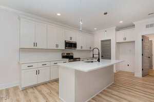 A modern kitchen with white cabinets, stainless steel appliances, a white island with a black faucet, light wood flooring, and two pendant lights hanging from the ceiling. Multiple gray doors are visible in the background.