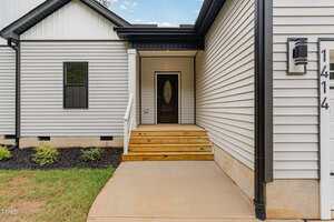 A concrete walkway leads to a wooden front porch with steps and a dark-colored front door. The house has light-colored siding, black trim, a window, and a house number sign reading 1414. Shrubs are planted along the side.