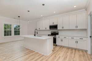 Modern kitchen with white cabinets, black hardware, stainless steel appliances, a large island with sink and black faucet, light wood flooring, white tile backsplash, and pendant lights above the island.