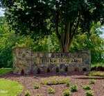A stone entrance sign reads "Lake Royale" at 100-106 Cherokee Drive, Louisburg, surrounded by a mulched area with small plants and flowers. A large tree and green foliage create a scenic backdrop under a partly cloudy sky.