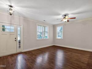 Empty living room with wood flooring, two ceiling lights, large windows with blinds, and a white front door. The room has light-colored walls and white trim. Natural light enters through the windows.