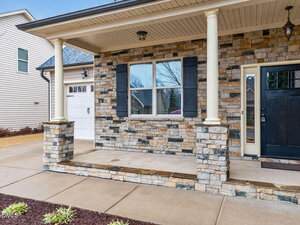 A small front porch with two columns, stone facade, black shutters, a black front door, and a window. The porch has a concrete floor and steps, with a driveway and siding visible on the left.