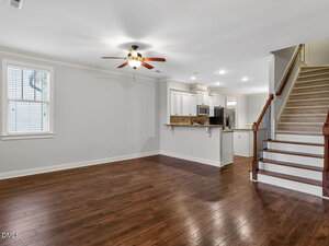 An empty living room with wood flooring, a ceiling fan, a window, and a view of the kitchen with bar seating. Carpeted stairs with a wooden railing lead to an upper floor on the right side.