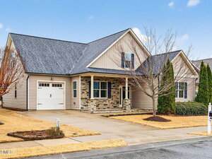 Single-story house with beige siding, brick accents, and a dark shingled roof. There is a front porch, attached single-car garage, and a driveway. The yard features minimal landscaping with small trees and shrubs.