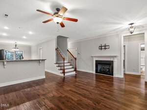 A spacious living area with dark wood floors, a ceiling fan, white walls, a fireplace, a staircase, and an open kitchen with a granite counter; natural light enters through windows in the background.