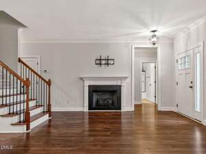 A spacious living room with hardwood floors, a white front door with glass panels, a black fireplace with a white mantle, light gray walls, and a staircase with wooden handrails and carpeted steps.