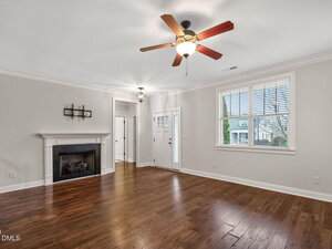 A spacious living room with hardwood floors, a ceiling fan, a white fireplace on the left wall, white walls, and large windows letting in natural light. A front door and hallway are visible in the background.