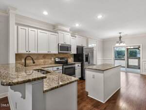 Modern kitchen with white cabinets, granite countertops, stainless steel appliances, tile backsplash, and wood flooring. A window and a glass door lead to a screened patio with backyard views. Ceiling lights and chandelier visible.