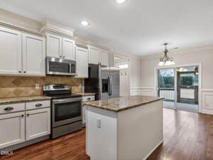A modern kitchen with white cabinets, stainless steel appliances, a granite island, and wood flooring. Sliding glass doors lead to an outdoor area, and a chandelier hangs above the adjacent dining space.