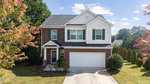 A charming two-story suburban house at 1320 Hosmer Court, Wake Forest, with brick and white siding exterior, green shutters, a two-car garage, and a landscaped front yard with trees and shrubs. The sky is mostly clear with a few clouds.