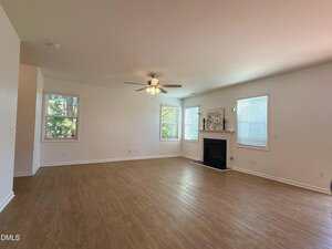 A spacious, empty living room with light-colored walls, wood flooring, three windows, a ceiling fan with lights, and a fireplace with a mantel and decorative artwork. Natural light fills the room.