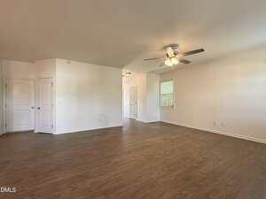 A spacious, empty room with light-colored walls, wood-style flooring, a ceiling fan with lights, and multiple closed white doors. A window with blinds lets in natural light from the right side.