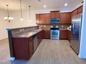 A spacious kitchen with wood cabinets, stainless steel appliances including a stove, microwave, and refrigerator, a double sink, dishwasher, brown countertops, and pendant lighting over a breakfast bar.