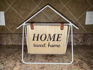 A wooden sign with the words "HOME sweet home" hangs on a white metal frame shaped like a house, set on a brown granite countertop against a tiled backsplash.