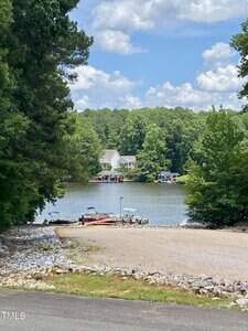 A small gravel boat ramp at 100 Cherokee Drive, Louisburg leads to a lake with several boats docked at the shore. Across the water, houses nestle among trees under a partly cloudy sky.