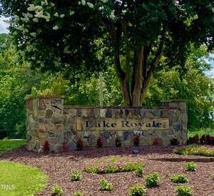 A stone sign with the text "Lake Royale" stands in front of a landscaped area with mulch, small plants, and a large tree in the background. The scene is outdoors with greenery and blue sky visible.