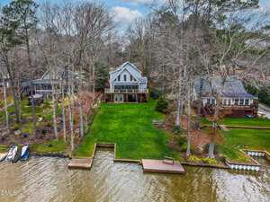 A two-story house with large windows and a deck overlooks a grassy lawn leading to a wooden dock on a lake. Neighboring houses and trees surround the property. The sky is partly cloudy.