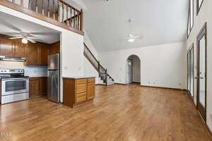 A spacious room with wood floors, an open kitchen featuring stainless steel appliances, wooden cabinets, a white countertop, and a loft area above. Stairs, ceiling fans, and large windows are visible.