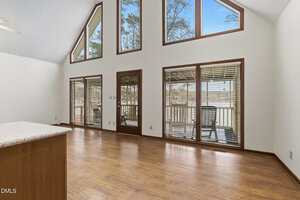 A spacious, empty room with wood flooring, large windows, and glass doors leading to a deck outside. The walls are white, and the ceiling is vaulted. Natural light fills the room through the windows.